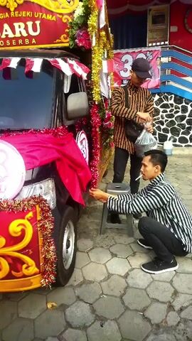 A video shows two men standing near a decorated car. The car is black and has a pink cloth draped over its front, along with red and gold streamers. One man, wearing a black and white striped shirt and black pants, is kneeling and appears to be adjusting the decorations on the car. He is holding a cigarette in his mouth. The other man, wearing a patterned shirt and a cap, is standing and holding a plastic bag. They are outdoors on a paved surface with a stone wall in the background. A banner with Indonesian text is visible on the vehicle.