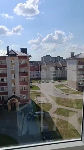 A view from a window looks out onto a street between apartment buildings under a blue sky with white clouds. In the foreground, a portion of the window frame is visible. The apartment building on the left is light tan and red with several floors and balconies. The building on the right is light tan and white, also with multiple floors and balconies. Across the street, more apartment buildings are visible, some with pitched roofs. A road runs between the buildings, with several cars parked or driving on it. Green grass and paved walkways are visible between the buildings and the road.