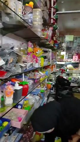 The video shows the interior of a cluttered shop. There are at least two women, one wearing a black hijab and the other wearing a light colored hijab. A boy is also present, visible from the back of his head. The women are browsing the shelves, which are stocked with various items including toys, decorative objects, and containers. One of the women takes a picture with her phone. The shop is brightly lit, and the shelves are packed with merchandise.