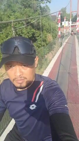 A man wearing a blue t-shirt, black cap, and sunglasses is riding a motorcycle across a suspension bridge. The bridge has red and white striped markings on its surface and is surrounded by green trees and a clear blue sky. Another motorcyclist is visible further ahead on the bridge. The video is a first-person perspective shot from the rider.