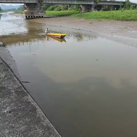 A person is in a yellow boat on a river. The boat is moving. The river has brown water. There is a bridge over the river in the background. There is a concrete embankment on the left side of the river. There is green vegetation on the right side of the river and under the bridge. The sky is overcast.