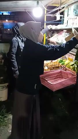 At night under a light a person wearing a gray hijab and a black jacket examines a bunch of green vegetables at a food stall.  Another person wearing a black hijab and a black jacket is standing to the left of the first person, watching. The food stall is covered with a white roof and has various food items on display. There is a pink plastic basket on the stall.  The area is dimly lit.