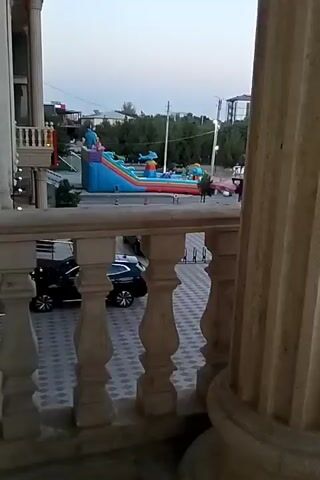 The video captures an outdoor scene with an inflatable playground, a large blue, green, and red bouncy castle with slides and various inflatable characters, set up in a paved area. To the left of the playground is a building with ornate columns and a balcony railing. Two black cars are parked in the foreground. In the background, there are trees, other buildings, and a utility pole. The sky appears overcast, suggesting it is either early morning or late afternoon.