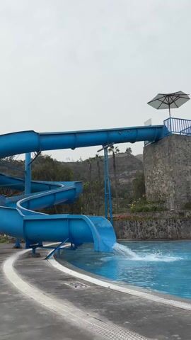A bright blue water slide stands next to a swimming pool. The slide is slightly damaged near the top. It is spiral shaped and ends by plunging into the water. A person descends the slide and makes a splash upon hitting the water. Palm trees and a rocky hill are visible in the background. A stone tower with a white umbrella above it is located behind the slide. The weather appears overcast.