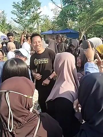 The video shows a group of people gathered outdoors, appearing to be at some kind of event or celebration. The central figure is a man wearing a black t-shirt with the words "Sahabat Rakyat" written on it. He is surrounded by people, mostly women wearing hijabs, who seem excited and cheerful. The man is smiling and interacting with the crowd. In the background, there are trees, a blue tent, and more people milling around. Some people are taking photos with their phones. The overall atmosphere is lively and festive.