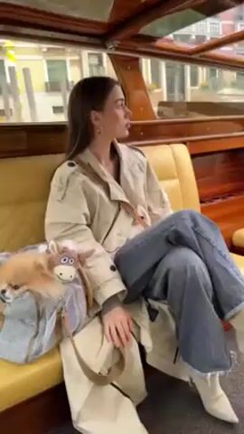 A woman with long brown hair sits on a yellow bench seat inside a boat. She is wearing a tan trench coat over a gray sweater and blue jeans with white boots. She has a purse slung over her shoulder. A small dog lies in a gray bag beside her. The boat is moving on a canal, as buildings can be seen through the windows. The interior of the boat is wood paneled and varnished.