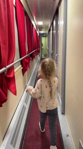 A small child with blonde hair braided in a single plait is seen from behind, walking down a narrow corridor inside a train. The corridor has red curtains hanging on one side, and gray-colored doors on the other. The child is wearing a light-colored sweater with a pattern of dogs on it, patterned gray pants, and white shoes. The floor of the corridor is covered with a red carpet. The lighting is bright and even. The child is moving away from the camera, walking towards a light at the end of the corridor.