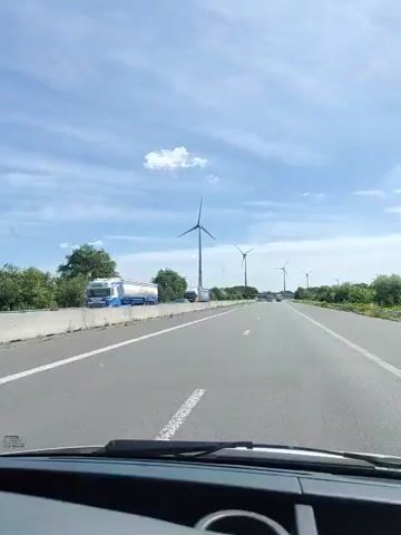 This video captures a first-person perspective from inside a vehicle traveling on a highway. The road ahead is a multi-lane highway with a concrete barrier on the left side. Several wind turbines are visible in the distance on the right side of the highway, interspersed with trees and vegetation. The sky is blue with scattered white clouds. Other vehicles are present on the highway, including a truck and several cars. The camera appears to be mounted on the dashboard of the vehicle, showing the windshield and the hood of the car. The video is in landscape orientation.
