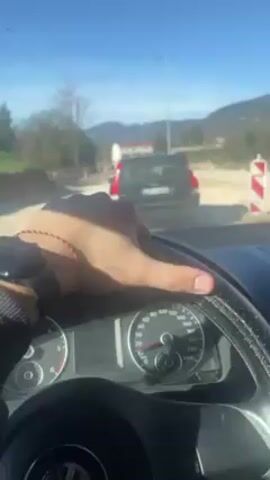 The video shows the interior of a car from the driver's perspective. The driver's left hand, wearing a dark smartwatch and a red and white bracelet, rests on the steering wheel. The car is moving at a slow speed as indicated by the speedometer. The car is behind a black SUV. Outside, the sky is clear and blue, and the surrounding environment consists of trees, mountains, and a building under construction, indicated by the presence of red and white traffic barriers. The car is moving forward slowly. The dashboard features a Volkswagen logo. The video is shaky.