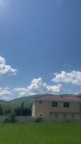 The video shows a two-story beige house with a red tile roof behind a green field. A small mountain or large hill is visible in the background, partially obscured by puffy white clouds in a blue sky. Trees are visible to the right of the house.