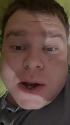 The video is a close-up, selfie-style recording of a man's face. He is lying down, with a green surface visible behind his head. The man's mouth is slightly open, and he appears to be speaking. His eyes shift from side to side, and he touches his forehead with his right index finger. The lighting is dim, and the man's face is illuminated from the front.