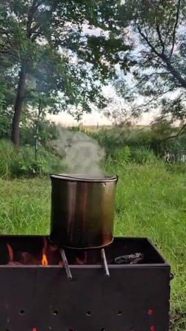 A pot of water is boiling over an open fire in a backyard. The pot is dark green and is sitting on two metal legs that are resting on a black barbecue grill. The flames from the fire are visible under the pot, and steam is rising from the pot. The backyard is filled with green grass and trees. The sky is cloudy and the sun is setting.