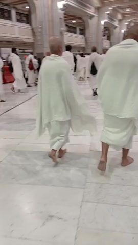 The video shows a group of pilgrims dressed in white ihram clothing walking barefoot in a large, ornate building. The floor is made of light-colored marble tiles, and the walls are decorated with arches and intricate designs. The pilgrims are walking in the same direction, and some of them are carrying bags. The lighting is bright, and the atmosphere appears calm and reverent.