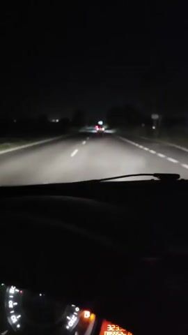 The video shows a first-person perspective of driving a car at night. The road has white lane markings and the headlights of the vehicle illuminate the dark surroundings. Several cars are visible ahead, their taillights glowing red. One car, in particular, is prominent in the frame, revealing its license plate which reads "B 6711 TT". A blue road sign is also visible to the right of this car. The overall environment appears to be a highway or a main road.