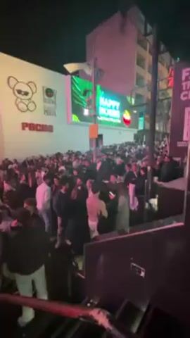 The video shows a large crowd of people gathered on a street at night. The crowd is dense and appears to be mostly young adults. Many are standing and talking, while some are looking at their phones. The environment includes buildings with neon signs and digital displays. One building has a large teddy bear graphic. The lighting is bright, with a mix of neon colors and streetlights illuminating the scene. The video seems to be taken from an elevated position, possibly a staircase or balcony.