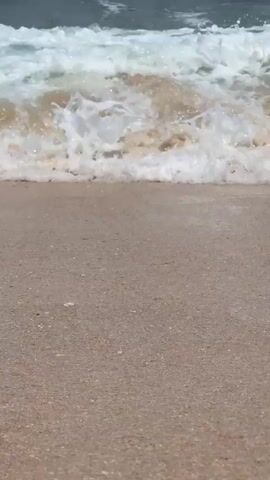 The camera focuses on a beach where waves are crashing onto the shore. The sand is a light tan color, and the waves are white and foamy as they reach the beach. The water further out is a darker gray-green color.