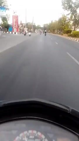 The video, captured from the perspective of a rider on a scooter, shows a street scene. In the foreground, a portion of the scooter's dashboard is visible, including a speedometer. The road ahead is paved, with painted lane markings. Several other motorcycles and scooters are traveling in the same direction. On the left side of the road, there appears to be a gas station with signage. Trees line the right side of the road. The sky is overcast or hazy. The time stamp indicates the video is from the very beginning of a recording, from 00:00 to 00:04.
