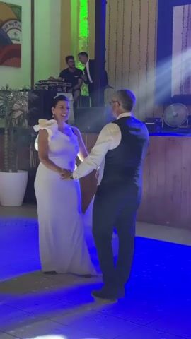 A wedding reception is underway, with a couple dancing in the center of the floor. The woman is wearing a white, one-shoulder dress with a flowing train, and the man is in a black vest over a white dress shirt and dark pants. They are holding hands and appear to be engaged in a romantic dance. In the background, a DJ booth is set up with two men standing behind it. One man is operating the DJ equipment, while the other appears to be a vocalist or MC. Another man in a suit is visible on a raised platform to the right of the DJ booth. The room is decorated with colored lights, including blue and green, which illuminate the dance floor. There are also some potted plants and a large fan in the background. The overall atmosphere seems festive and celebratory.