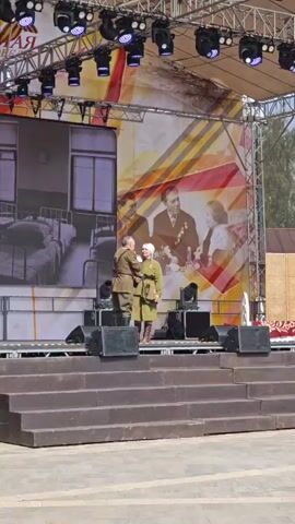 The video shows a staged reenactment or performance taking place on an outdoor stage. Two individuals, dressed in what appears to be military or historical-themed costumes, stand on the stage. The person on the left seems to be pinning or adjusting something on the uniform of the person on the right.

The stage is constructed with a series of tiered steps leading up to a platform. Behind the performers, there is a large backdrop. The backdrop features a black and white image of a hospital ward on the left side, and a colored image of people on the right.

The stage is equipped with lighting fixtures mounted on a metal frame above. Several black speakers are positioned on the stage floor. To the right of the performers is a partition screen.

The setting appears to be a town square or public space, based on the surrounding buildings. The sky is visible in the background, and it seems to be a partly cloudy day.