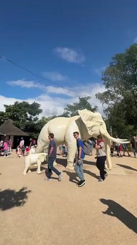 A large, white, elephant statue is displayed outdoors on a sunny day. The statue is accompanied by a smaller, white, baby elephant statue. Several people are walking around the statues. The ground is dirt and gravel. There are trees and a building with a thatched roof in the background. The sky is blue with scattered white clouds.