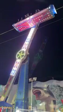 At night, the video shows a carnival ride in operation.  The ride is a pendulum style, with a long arm that swings back and forth, carrying a platform of people at its end. The arm is lit with white and colored lights. The platform is red and also has white lights.  The base of the ride is blue and decorated with lights as well. In the background, there is a large painted backdrop featuring a shark jumping out of the water.  A few people are seen walking on the ground, appearing as spectators. The overall scene is colorful and lively.