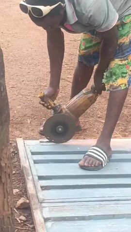 A man in flip-flops, sunglasses, and a short-sleeved shirt and shorts is cutting a piece of grey metal with a powered angle grinder. He appears to be outside on a dirt-covered ground. He is holding the grinder with both hands and carefully cutting through the metal, creating sparks. A tree and other objects are visible in the background.