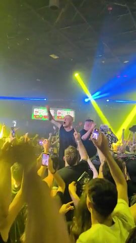 A video shows a concert in a dimly lit venue. Two bald men stand on a stage, facing a crowd of people. The man on the left is singing and gesturing with his arms. The man on the right is holding a phone up to record. Both men are wearing dark t-shirts. The crowd is cheering and holding up their phones. Spotlights are shining on the stage. The venue appears to be a club or concert hall with a high ceiling and banners in the background.