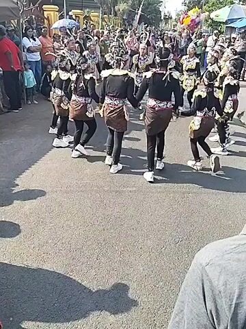The video shows a group of approximately ten young performers in matching traditional attire, consisting of black tops, brown patterned skirts, and headwear, dancing in a circular formation on a paved road. They are holding hands and moving in unison, with some bending their knees and swaying. A crowd of spectators, including adults and children, is gathered on both sides of the road, watching the performance. The environment appears to be outdoors, with buildings and trees visible in the background. The lighting suggests it is daytime.