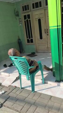 The video shows an elderly man sitting in a turquoise plastic chair outside a house. He is asleep, with his head tilted back and his body leaning against the chair. He is wearing a striped shirt and shorts. The house appears to be a modest dwelling with a green exterior and a white door. There is a tiled porch area with some shoes and an umbrella visible. The sun is shining, casting shadows on the ground. The overall scene is one of peaceful repose in a residential setting.
