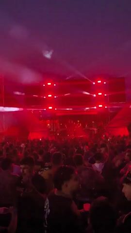 The video shows a crowded outdoor music festival at night. The stage is lit with red and white lights, and there are large screens displaying abstract imagery. A DJ booth is visible on stage. The crowd is diverse, with people of various ages and ethnicities dancing and enjoying the music. Some individuals are holding drinks, and others are recording the event on their phones. The atmosphere appears energetic and celebratory.