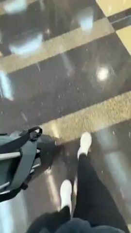 A person wearing black pants and white shoes walks across a tile floor with a piece of luggage. The tile floor has a black and tan checkerboard pattern. The luggage is dark gray and black with silver accents. The person is walking towards an indeterminate location.