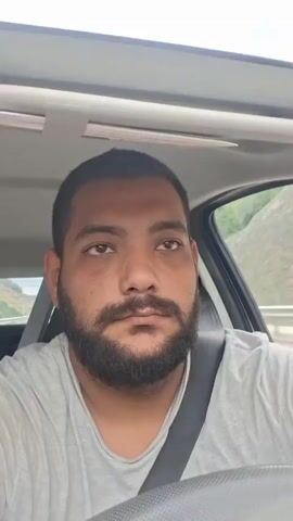 The media file shows a selfie video of a man with a beard, wearing a grey t-shirt and a seatbelt, sitting in the driver's seat of a car. The man is looking directly at the camera and appears to be speaking or singing, with his mouth open. The scene is set inside a car, with the dashboard and part of the steering wheel visible in the foreground. The background shows a glimpse of the outside through the car window, suggesting the vehicle is in motion, possibly on a road with a hillside and some vegetation. The lighting suggests it is daytime. No visible text is present in the media file.