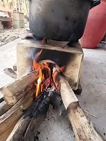 A close-up shot focuses on a wood-burning stove with a large black pot on top. The fire is actively burning, with flames licking up from the logs placed in the stove. The stove appears to be made of concrete or clay. The ground around the stove is concrete. In the background, there are some indistinct objects and structures, possibly indicating an outdoor setting. A large red object, possibly a barrel or container, is visible on the right side of the frame. The overall scene depicts a traditional method of cooking.