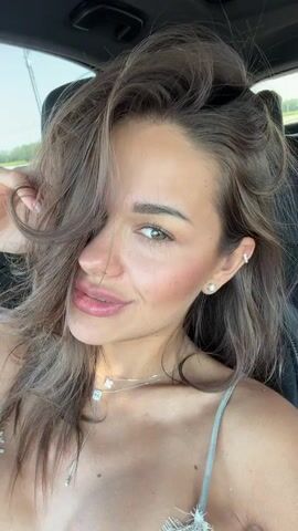 The video shows a close-up, selfie-style shot of a young woman sitting in a car. She has long, wavy brown hair that partially covers her face. She is wearing light-colored spaghetti strap clothing and a necklace with several small pendants. She has earrings in both ears, including a small stud in her left ear and a larger, more decorative earring in her right ear. She is looking at the camera and smiling, with her right hand touching her hair. The background is blurred, but appears to be outdoors with some greenery visible. The lighting suggests it is daytime.