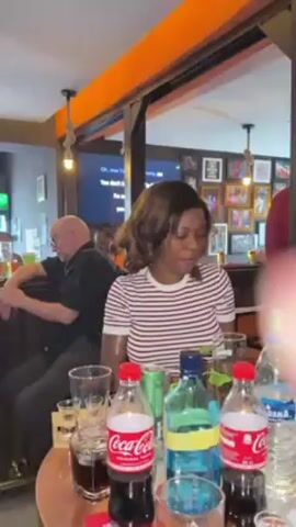 The video shows a woman standing behind a bar, in what appears to be a pub or a similar type of establishment. She has brown hair and is wearing a black and white striped shirt. In front of her on the bar are bottles of Coca-Cola, a blue alcoholic beverage, other bottles and glasses, some with drinks in them. In the background, other patrons are visible at the bar, including an older man in a black shirt and glasses sitting at the bar. There is a television screen showing lyrics, and several framed pictures on the back wall. The lighting in the pub is moderate, with a combination of natural and artificial light. The woman is looking at the camera, speaking and gesticulating.