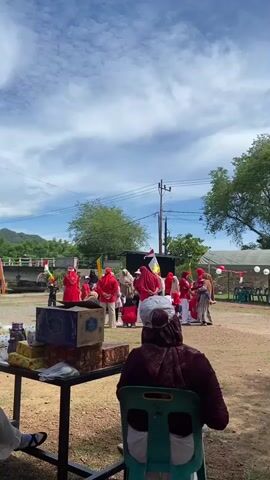 A group of people, mostly wearing red and white clothing, are gathered outdoors under a cloudy sky. Some individuals are wearing headscarves. There is a table in the foreground with boxes on it. In the background, there are trees, a utility pole, and a tent. Some people appear to be watching or participating in an event.