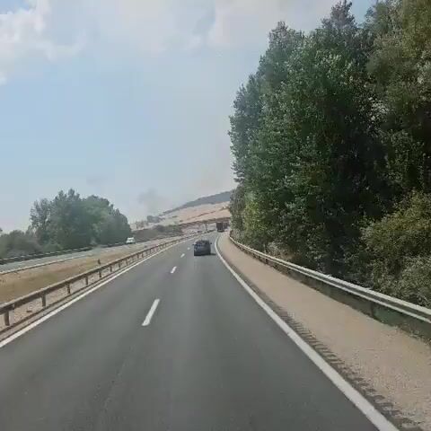 A highway scene on a clear day, viewed from a vehicle. The road has multiple lanes separated by white lines and a guardrail on the right. Trees line the right side of the road, and a hill with dry, golden vegetation is visible in the distance. Several cars and a truck are on the highway. Smoke plumes rise from the distant hill. A pedestrian overpass spans the highway in the middle ground.