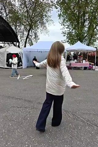 The video shows a woman and a man in an outdoor setting with vendor tents in the background. The woman is wearing a white padded jacket and a long blue skirt. She is smiling and twirling, with her arms outstretched. The man is wearing a historical-style tunic and pants. He is also twirling and smiling. There are several tents set up, and a few people can be seen milling around them. The ground is paved, and there are some trees in the background. There appears to be music playing.