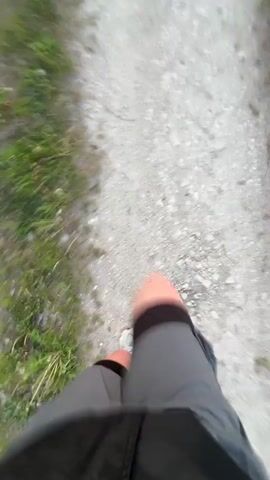 A person is running on a dirt path. The camera is positioned to show the person's legs and the path ahead. The path is flanked by green vegetation on either side. As the person continues running, the camera angle shifts, and the view opens up to reveal a scenic sunset at the end of the path. The sun appears as a bright orange orb, silhouetting the trees and vegetation along the path. The sky transitions from a light blue near the horizon to a darker shade above.