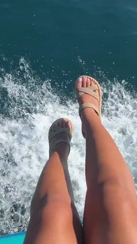 The video begins with a close-up of a woman's legs and feet as she sits with her legs dangling over the side of a boat. She is wearing tan sandals with a braided strap and has painted toenails. The water is a deep blue and is creating white foam and spray as the boat moves. The camera then pans up to reveal a coastline with green, tree-covered mountains and a few buildings. The sun is shining and the water is calm.