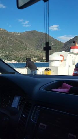 The view from inside a car on a ferry shows a person leaning on the ferry's railing, looking out at the water and mountains. The person is wearing dark clothing. Part of the ferry’s structure is visible. In the foreground, the car's dashboard and a hanging cross are visible. The weather appears sunny with a clear sky.