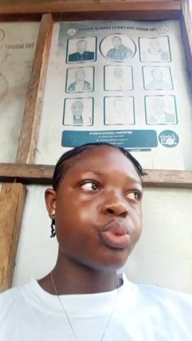 A young woman with dark skin and closely braided hair is the central subject of the video, filmed in a close-up, chest-up shot. She wears a plain white short-sleeved t-shirt and a silver chain with a shiny, ornate pendant around her neck. A small stud earring is visible in her left ear, and her fingernails are painted black. Her skin appears slightly shiny, suggesting warmth or humidity.

Throughout the video, she exhibits varying expressions: initially pouting and looking off to her right, then closing her eyes momentarily, and finally looking towards the camera while making several hand gestures. Her right hand first comes up with the index finger to her lips, then points downwards, and finally moves in a quick, dismissive gesture.

The background features a large, rectangular poster or sign mounted on a rustic wooden frame. The text on the poster, which appears upside down, clearly reads "THE MOUNT ZION CHURCH NIGERIA" and "IMPORTANT CHURCH EVENTS." Below the main title, nine framed portraits of men, presumably clergy or significant figures, are displayed in a grid pattern. A small logo at the bottom right of the poster (also upside down) shows "Zion 2021."

The bright lighting indicates it is daytime, likely sunny. The wooden structure framing the poster suggests an open-air or semi-enclosed setting, possibly a public space or simple shelter in Mbribit, Nigeria.