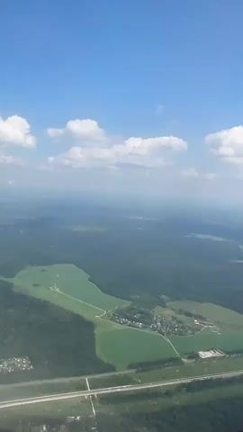 This is a first-person perspective video taken from an airplane window, looking down at a rural landscape. The scene is set under a clear blue sky with scattered white cumulus clouds. The time of day appears to be daytime, likely late morning or early afternoon, given the brightness of the sun.

The dominant feature of the landscape is a vast expanse of lush green forests, interspersed with lighter green agricultural fields. A prominent feature is a large, irregularly shaped green field that resembles a lake or a specially cultivated area. Within this field, there are some visible structures, possibly farm buildings or houses. A small cluster of buildings, resembling a village or hamlet, is situated near the edge of this large green area, with roads connecting it to other parts of the landscape. In the distance, the landscape appears hazy, suggesting a considerable distance and possibly some atmospheric conditions.

The video also shows a glimpse of the airplane's window frame, indicating the vantage point. The overall mood conveyed is one of tranquility and vastness, typical of aerial views of natural landscapes. There is no visible text in the video. The location context of Borduki, Russia, suggests the area being viewed is in that region.