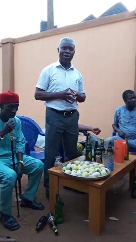 A man in a light-colored shirt and gray pants is standing and speaking to three other men. One of the men is seated to the left wearing a light blue outfit with a red hat and holding a cane. The other two men are seated to the right, wearing blue shirts. A wooden table is in the center, covered with bottles of beer and a silver tray full of white round objects, possibly eggs. A plastic orange pitcher and clear glasses are also on the table. The scene appears to be outdoors in front of a beige wall. The speaker is gesturing with his hands while addressing the other men.