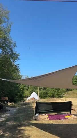 The video shows a clear blue sky with a small helicopter flying in the distance. In the foreground, there is a makeshift shelter with a large tan tarp overhead and a black tarp covering a low structure. Several blankets or sleeping bags are visible on the ground near the shelter. Trees and foliage surround the area, and a white tent or structure is visible further in the background. The ground appears to be dry grass and dirt. A wire is strung across the top of the frame. A voice in Russian can be heard saying, "And the phone is charging."