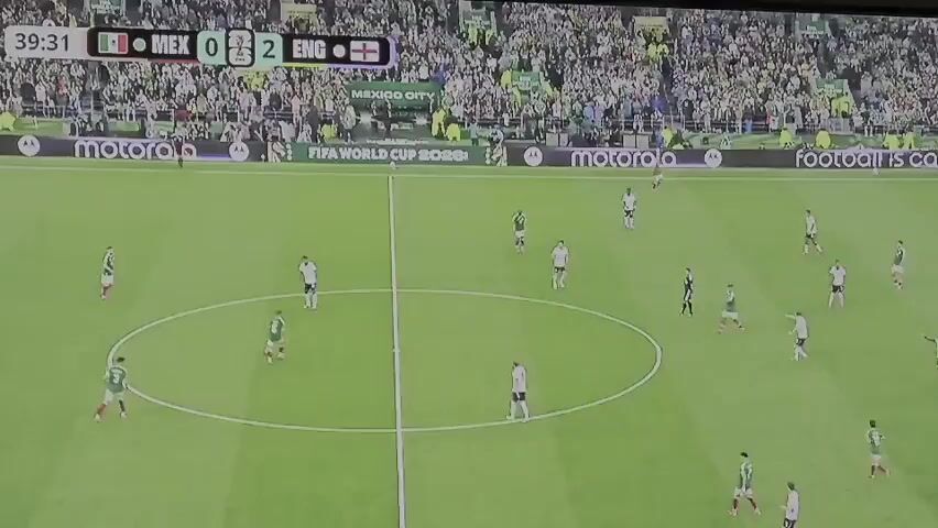 This is a wide shot of a soccer game. A scoreboard at the top of the screen shows Mexico with 0 points and England with 2 points. The time is 39:32. The field is green grass with white lines. There are many players on the field, wearing green and white jerseys and white and black jerseys. The players in green and white are Mexico, and the players in white and black are England. There is a crowd of spectators in the background. The spectators are wearing a variety of clothing. There are several advertisements visible around the field. The players are spread out across the field. One player in a green and white jersey is kicking a ball in the center of the field. The referee is standing near the center circle.