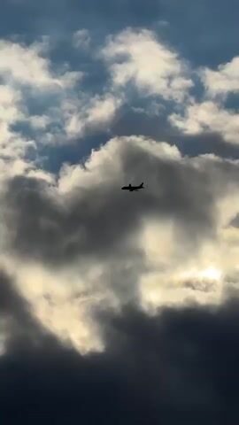 The media file shows a silhouette of a passenger airplane flying through a cloudy sky. The plane is dark against the lighter clouds, which are illuminated by sunlight filtering through them. The clouds appear dense and textured, with some areas of bright white and others of dark gray. The sky above is a deep blue, hinting at a clear patch of sky further up. The overall impression is of a dramatic and somewhat moody sky.

The activity taking place is simply the flight of an airplane. There are no people or specific objects visible besides the aircraft. The scene is set in the sky, with no discernible ground features. The time of day is not explicitly stated but the lighting suggests it could be daytime, possibly late afternoon or early morning, given the interplay of light and shadow on the clouds. The weather appears to be mixed, with both bright and dark clouds present.

There is no visible text in the media file. The location context provided is Пычанки, Russia, which is a geographical place, but no visual cues within the image directly confirm this location. The emotions evoked by the image could be awe, wonder, or a sense of adventure, due to the majestic presence of the airplane against the dramatic sky.