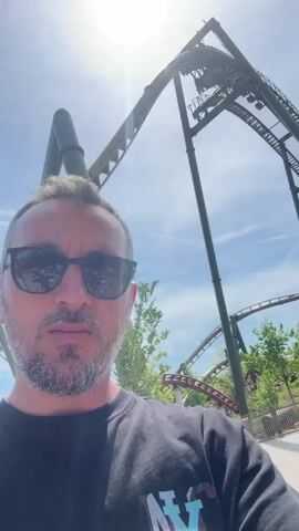 The video presents a first-person view of a man wearing sunglasses and a dark t-shirt standing in what appears to be a theme park. In the background, a roller coaster with a green track is visible against a partly cloudy sky. The roller coaster cars are seen moving along the track. Other people can be seen walking in the background, and there are elements of the park's scenery, including greenery and wooden structures. The man remains still, looking at the camera, while the roller coaster operates behind him.