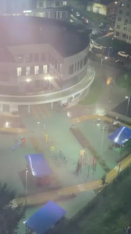 This aerial video shows a playground and a parking lot at night during a rainstorm. The playground is empty, with swings and slides visible. The parking lot is filled with parked cars. The rain is heavy, and the ground is wet and reflective. The video appears to have been taken from a high vantage point, looking down on the scene.