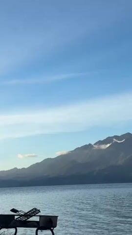 The video shows a wide shot of a tranquil lake with mountains in the background. The sky is a clear blue with a few wispy clouds. In the foreground, a dark object, possibly a grill, is partially visible on the left side of the frame. The water is calm, reflecting the light blue of the sky. The mountains are a deep green and appear to be covered in lush vegetation, with some patches of white clouds clinging to their peaks. The overall atmosphere of the video is peaceful and serene, suggesting a quiet moment in nature. There are no people or animals visible in the frame. The lighting suggests it is daytime, possibly early morning or late afternoon, given the soft quality of the light.